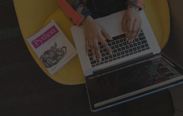 A person typing on a laptop placed on their lap, seated on a bright yellow chair. A Python programming book is positioned next to the laptop, with a second screen showing lines of code.