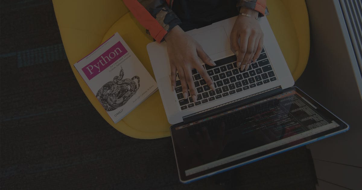 A person typing on a laptop placed on their lap, seated on a bright yellow chair. A Python programming book is positioned next to the laptop, with a second screen showing lines of code.
