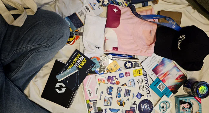 A collection of swag from WordCamp Asia 2025 spread out on a bed, including stickers, notebooks, t-shirts, a lanyard, a cap, and other event freebies.