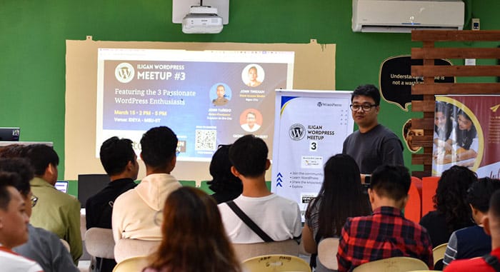 Audience at Iligan WordPress Meetup #3 listening to a speaker, with an event slide on screen showing featured speakers and event details.