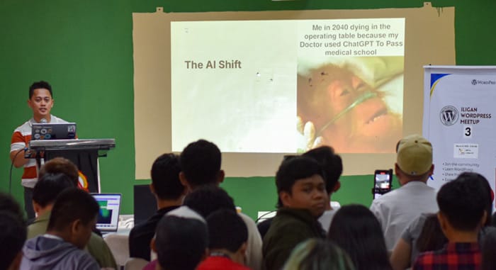 John is presenting at a WordPress Meetup in Iligan, discussing “The AI Shift” with a humorous slide referencing ChatGPT and medical school. Audience seated and engaged.