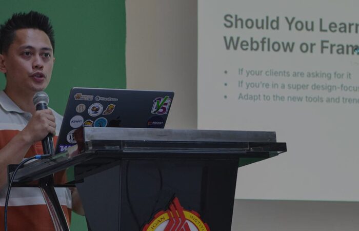 Me giving a talk at a WordPress event, standing at a podium with a laptop covered in tech stickers. A slide behind him reads, “Should You Learn Webflow or Framer?