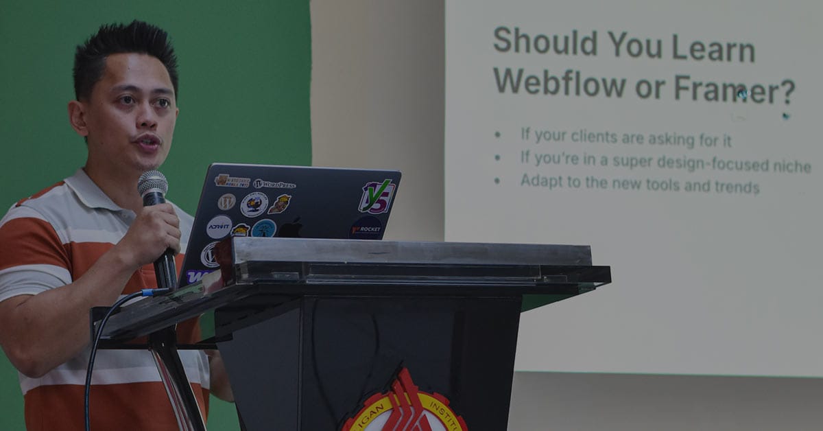 Me giving a talk at a WordPress event, standing at a podium with a laptop covered in tech stickers. A slide behind him reads, “Should You Learn Webflow or Framer?