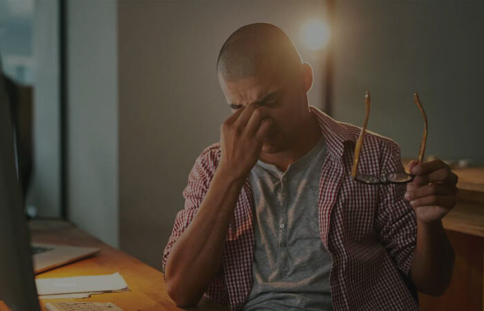 A stressed freelancer feeling anxious about his job security, sitting at his desk with his glasses off while pinching the bridge of his nose.