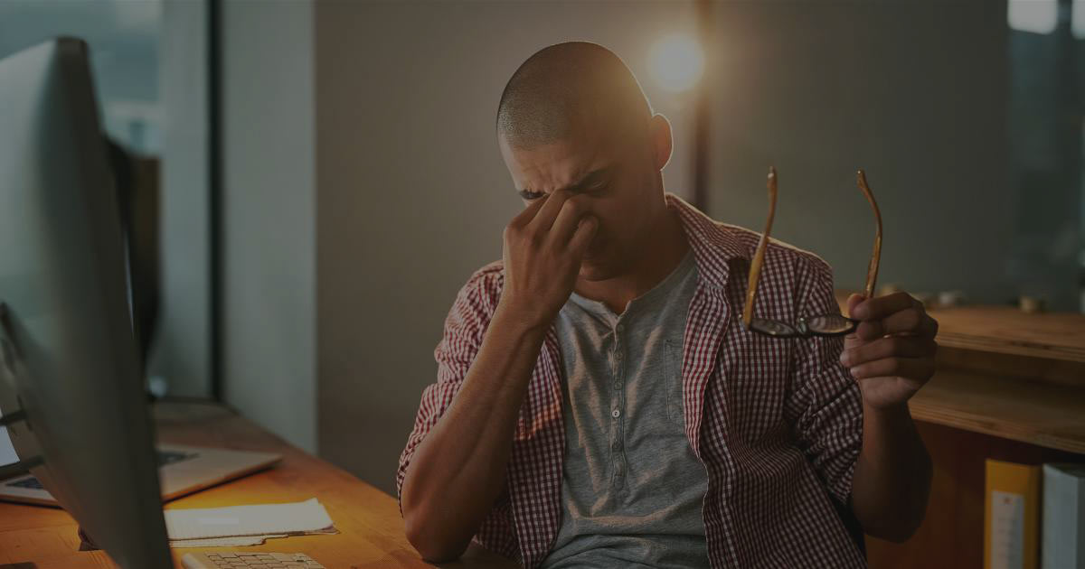 A stressed freelancer feeling anxious about his job security, sitting at his desk with his glasses off while pinching the bridge of his nose.