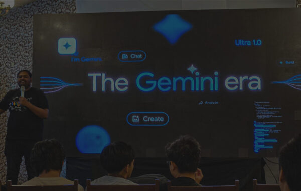 An image from a Google I/O Extended event, showing a speaker standing on a stage. The screen behind him features the phrase "The Gemini era" and several interactive icons, while an audience watches from the front rows.