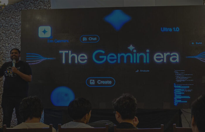 An image from a Google I/O Extended event, showing a speaker standing on a stage. The screen behind him features the phrase "The Gemini era" and several interactive icons, while an audience watches from the front rows.