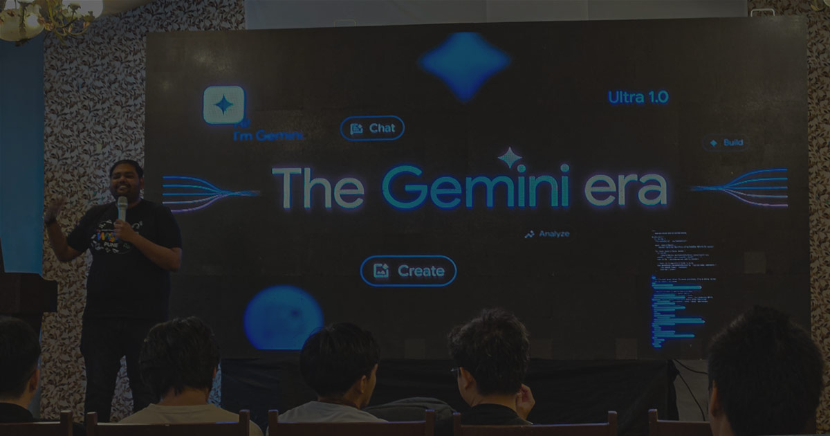 An image from a Google I/O Extended event, showing a speaker standing on a stage. The screen behind him features the phrase "The Gemini era" and several interactive icons, while an audience watches from the front rows.