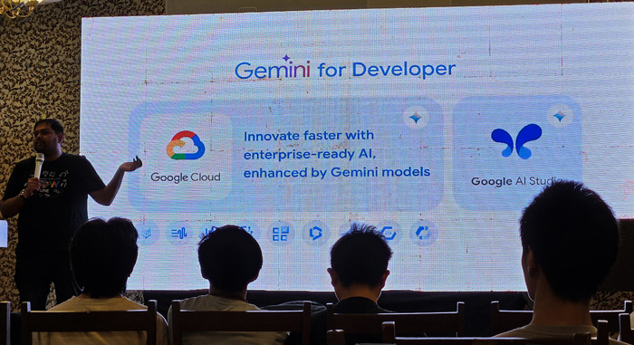 Vrijraj Singh is on a stage, speaking and gesturing to a large screen. The screen displays a presentation slide titled "Gemini for Developer" and showcases Google Cloud and Google AI Studio as tools for innovating faster with enterprise-ready AI.
